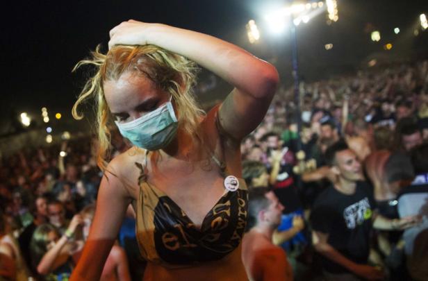 Eine junge Frau mit Maske und ungewöhnlichem Oberteil steht vor einer Menschenmenge.