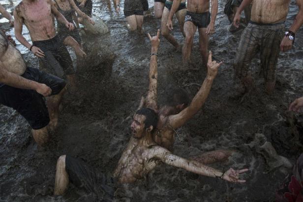Mehrere Menschen, mit Schlamm bedeckt, stehen und liegen in einem schlammigen Bereich und zeigen Handgesten.