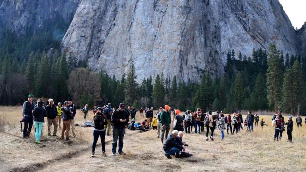 Eine große Gruppe von Menschen steht auf einer Wiese vor einem hohen Felsen.