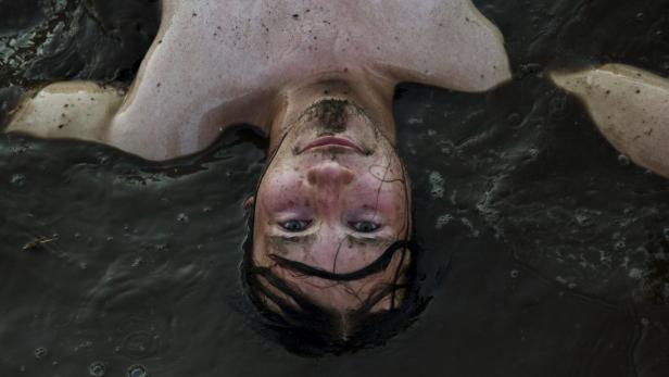 Ein Mann liegt im trüben Wasser, sein Gesicht und Oberkörper sind mit Schlamm bedeckt.