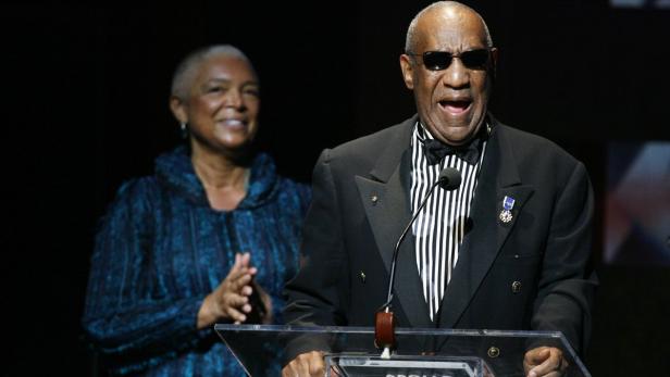 Bill Cosby spricht am Rednerpult, während eine Frau im Hintergrund applaudiert.