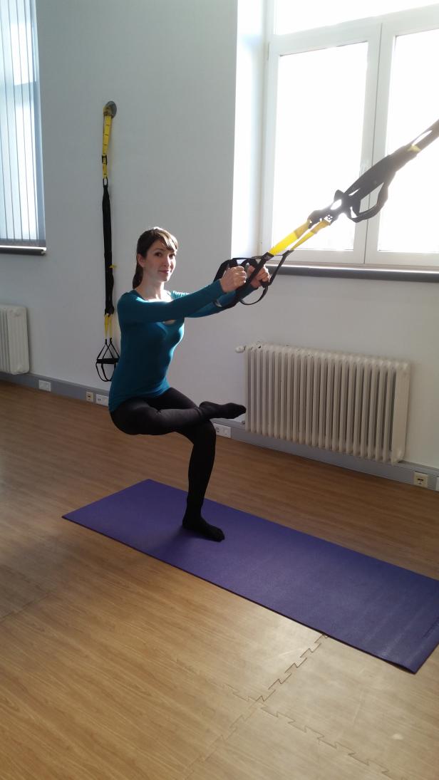 Eine Frau macht Kniebeugen mit einem gelben TRX-Band auf einer lila Matte.
