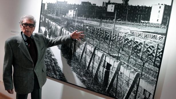 Ein älterer Mann steht vor einem Schwarzweißfoto der Berliner Mauer.