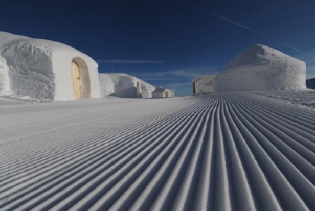 Schnee bedeckte Iglu-ähnliche Strukturen auf einer Skipiste unter blauem Himmel.