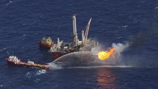 Eine Ölplattform auf dem Meer wird mit Wasser bespritzt, während eine Stichflamme aufsteigt.
