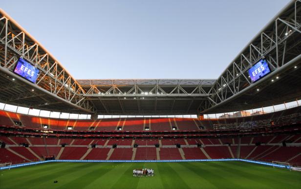 Blick in ein Fußballstadion mit roten Sitzen und einer Gruppe von Spielern auf dem Rasen.