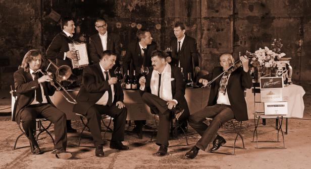 Eine Gruppe Musiker in Anzügen posiert mit Musikinstrumenten und Weingläsern vor einer Mauer.