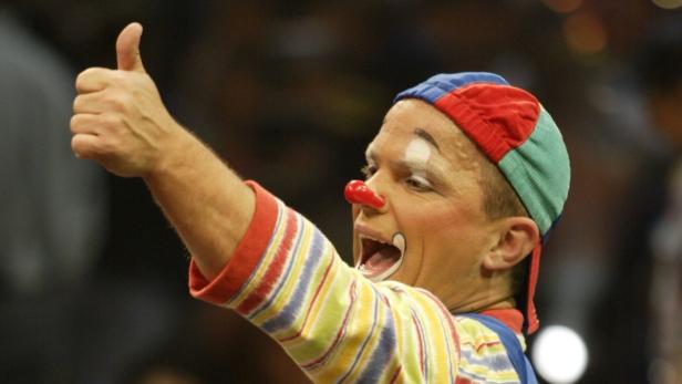 Ein Clown mit roter Nase und bunter Kleidung zeigt den Daumen nach oben.