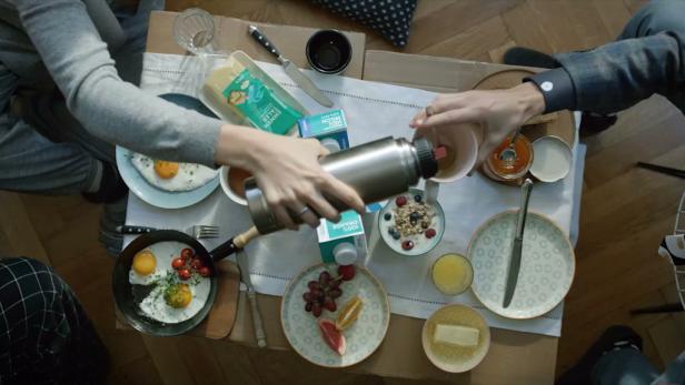 Vogelperspektive auf einen Frühstückstisch mit Eiern, Müsli, Obst und einer Person, die Kaffee einschenkt.