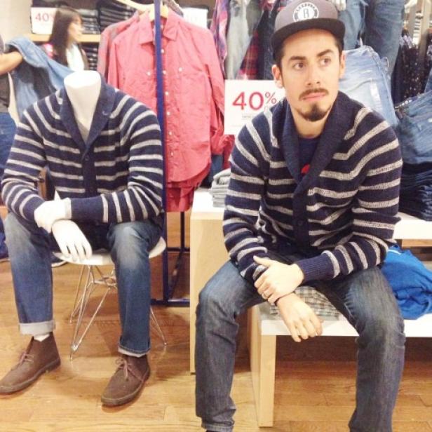 Ein Mann mit Mütze sitzt neben einer Schaufensterpuppe im gleichen gestreiften Cardigan.