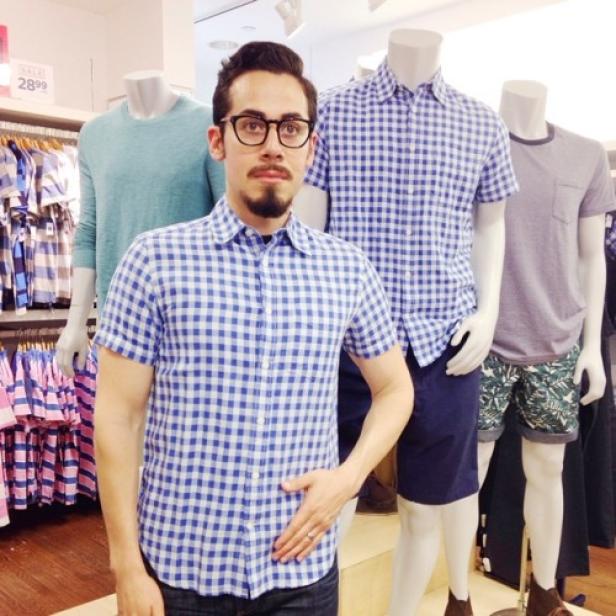 Ein Mann mit Brille und kariertem Hemd posiert in einem Bekleidungsgeschäft neben Schaufensterpuppen.