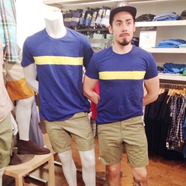 Ein Mann posiert neben einer Schaufensterpuppe, beide tragen ein blaues T-Shirt mit gelbem Streifen und Khaki-Shorts.