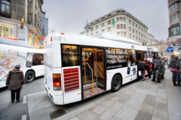Ein weißer Bus hält an einer Haltestelle in der Stadt, Fahrgäste steigen ein und aus.