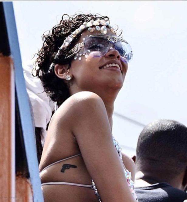 Rihanna lächelt mit Schmuck, Sonnenbrille und einem Pistolen-Tattoo auf der Haut.