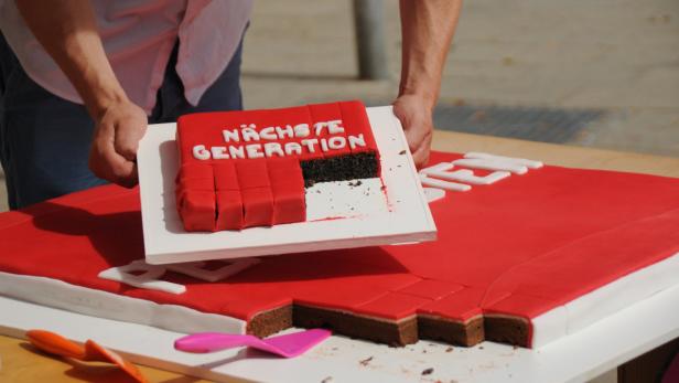 Eine rote Torte mit der Aufschrift „Nächste Generation“ wird angeschnitten.