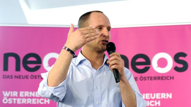 Ein Mann spricht vor einem pinkfarbenen Hintergrund mit dem Logo der Partei NEOS.