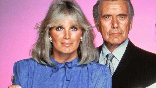 Eine Frau mit blonden Haaren und ein Mann im Anzug vor einem rosa Hintergrund.