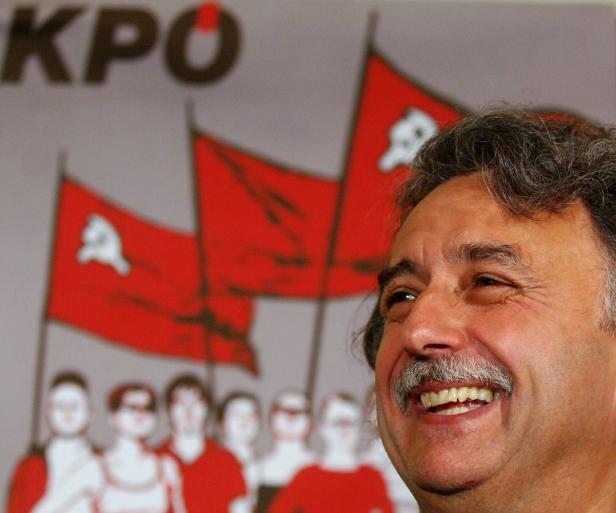 Ein lächelnder Mann steht vor einem Hintergrund mit KPO-Logo und roten Fahnen.