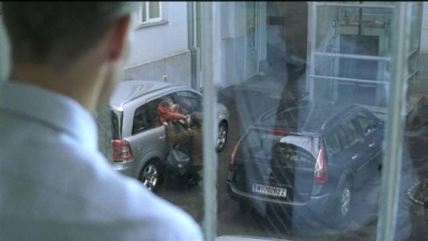 Ein Mann blickt aus dem Fenster auf zwei Autos und Personen, die Gepäck einladen.