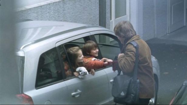 Ein Mann verabschiedet sich von zwei Kindern, die mit einem Stofftier im Auto sitzen.