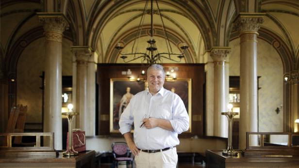 Ein Mann steht in einem eleganten Raum mit Säulen und einem Gemälde im Hintergrund.