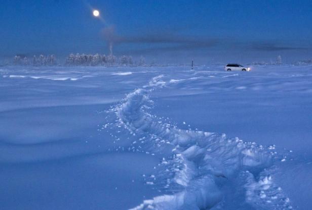 Eine Spur im Schnee führt zu einem Auto in einer verschneiten Landschaft unter einem Mondschein.