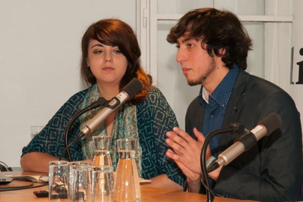 Eine Frau und ein Mann sitzen an einem Tisch mit Mikrofonen und Gläsern.