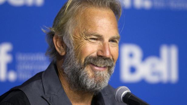 Kevin Costner lächelt bei einer Pressekonferenz in ein Mikrofon.