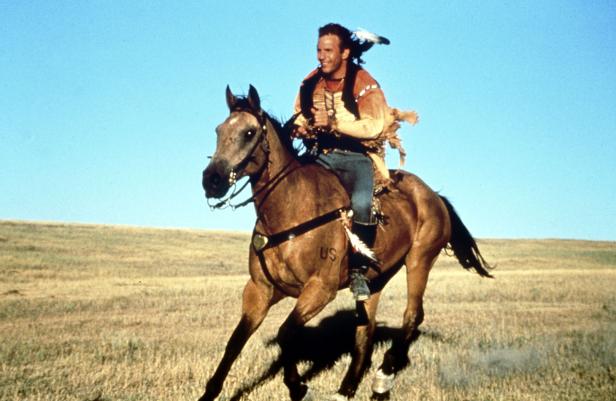 Ein Mann mit indianischer Kleidung reitet auf einem Pferd über eine weite Ebene.