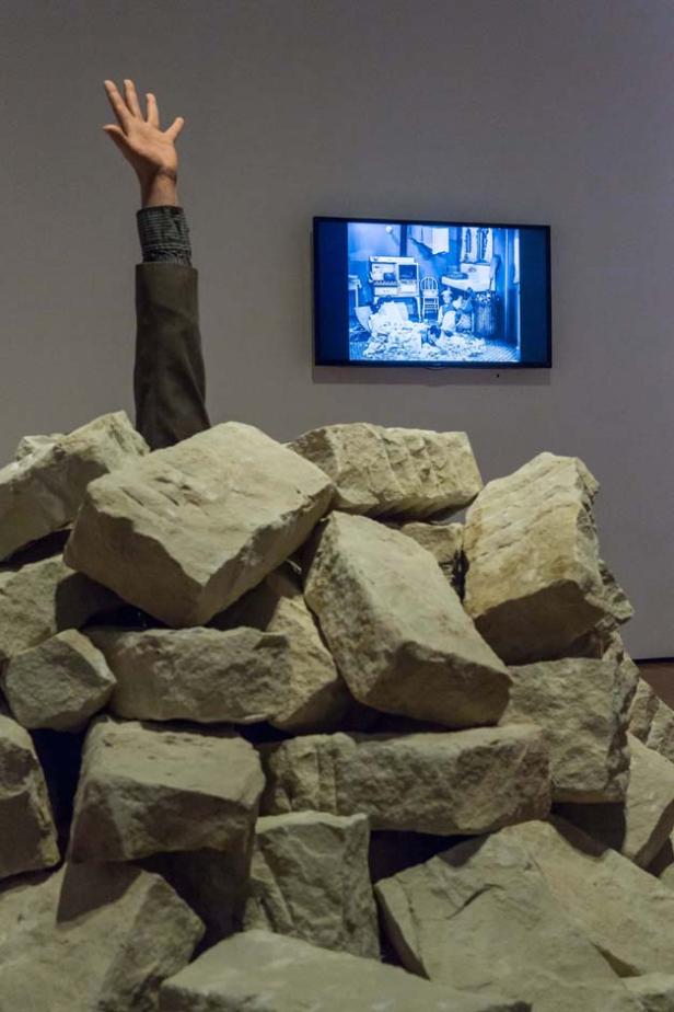 Ein Arm ragt aus einem Steinhaufen, im Hintergrund ein laufender Fernseher.