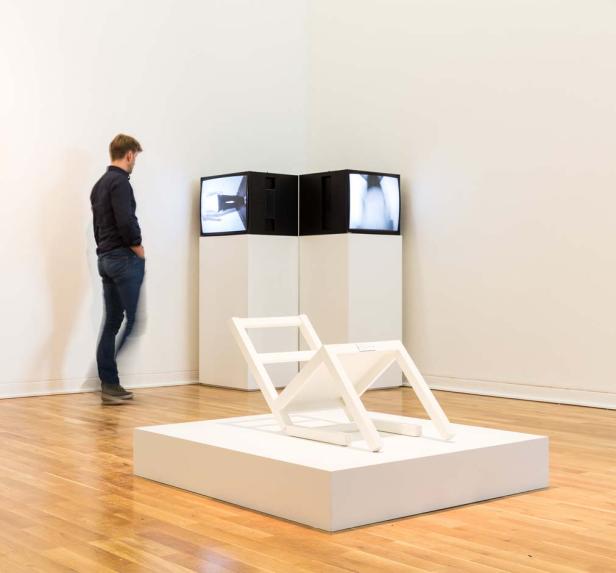 Ein Mann betrachtet eine Kunstinstallation mit einem weißen Stuhl und zwei Monitoren.