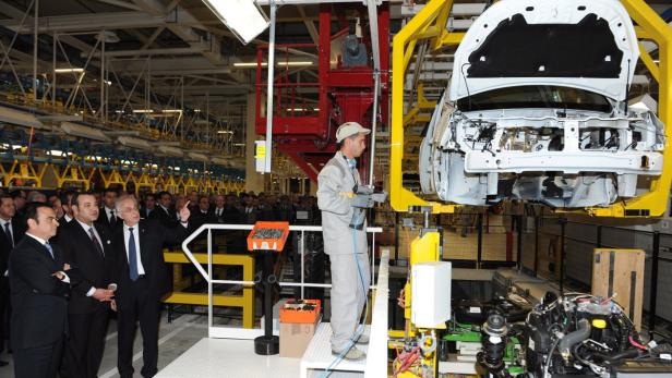 Ein Arbeiter montiert in einer Fabrik ein Auto, während eine Gruppe von Anzugträgern zusieht.
