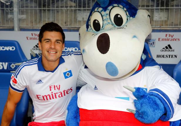 Ein lächelnder Mann im HSV-Trikot posiert mit dem Vereinsmaskottchen Dino Hermann.