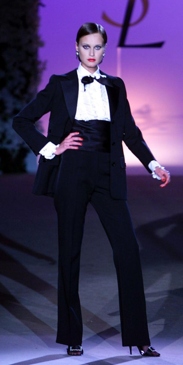 Ein Model präsentiert einen schwarzen Hosenanzug mit Rüschenbluse auf dem Laufsteg.