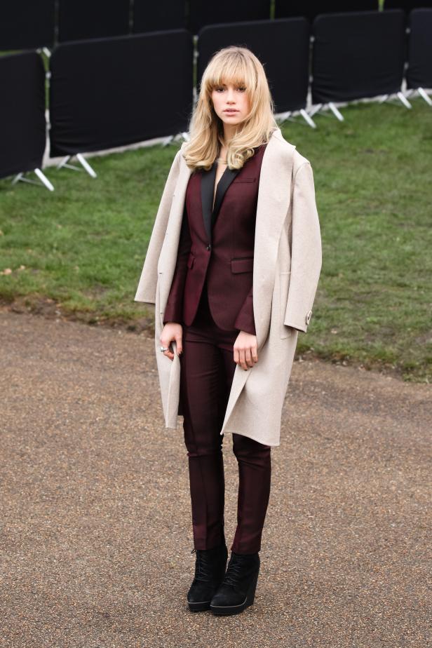 Eine Frau in einem burgunderfarbenen Hosenanzug und einem beigen Mantel steht im Freien.