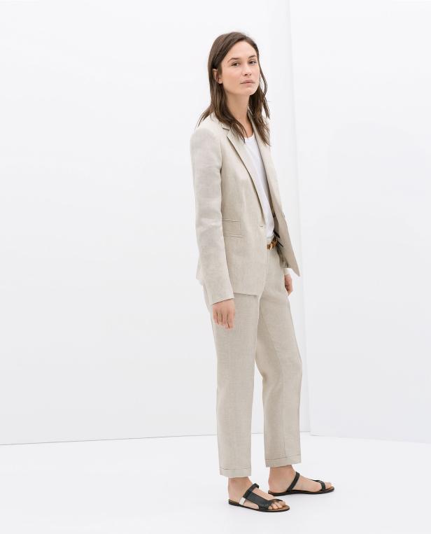 Eine Frau in einem hellen Hosenanzug und Sandalen steht vor einer weißen Wand.