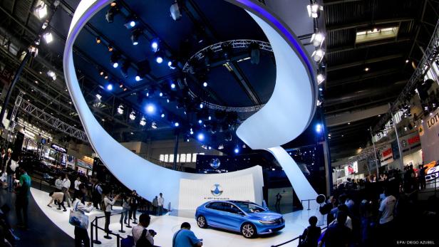 Ein blauer Lenza auf einer Automesse unter einer großen, geschwungenen Lichtinstallation.