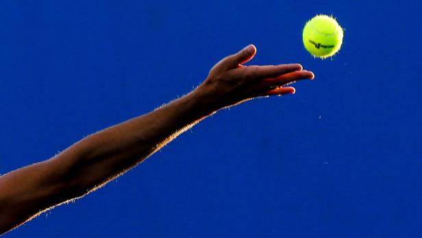 Ein Arm wirft einen Tennisball vor einem blauen Hintergrund in die Luft.