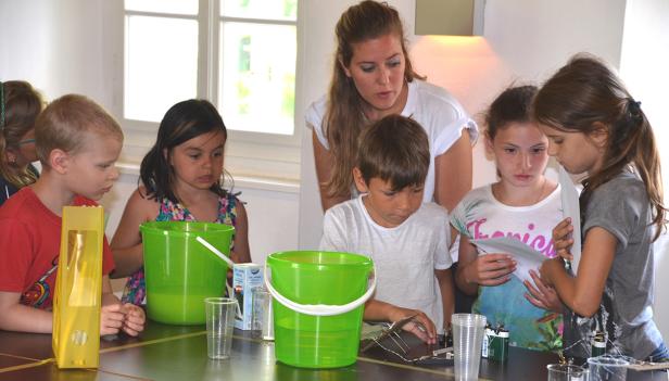 Eine Gruppe Kinder experimentiert unter Aufsicht einer Frau mit Flüssigkeiten und elektrischen Bauteilen.