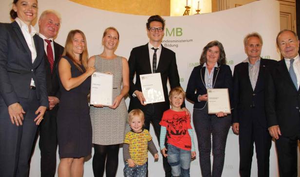 Eine Gruppe von Erwachsenen und zwei Kinder posieren mit Urkunden vor einer Wand des Bundesministeriums für Bildung.