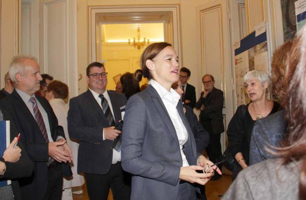 Eine Frau in einem grauen Hosenanzug steht in einer Gruppe von Menschen in einem eleganten Raum.
