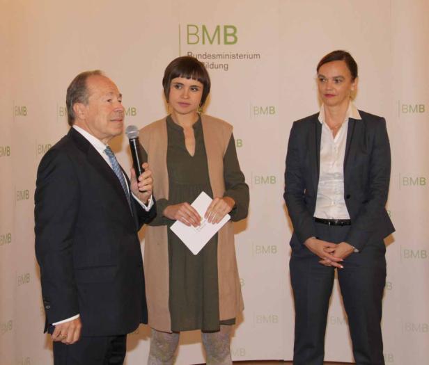 Drei Personen stehen vor einem Hintergrund mit dem Logo des Bundesministeriums für Bildung (BMB).