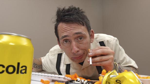 Ein Mann hält eine Zigarette in der Hand, umgeben von Cola, Chips und einem Aschenbecher.