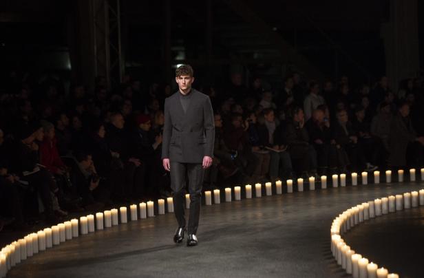 Ein Mann in einem grauen Anzug läuft über einen Laufsteg, der von Kerzen gesäumt ist.