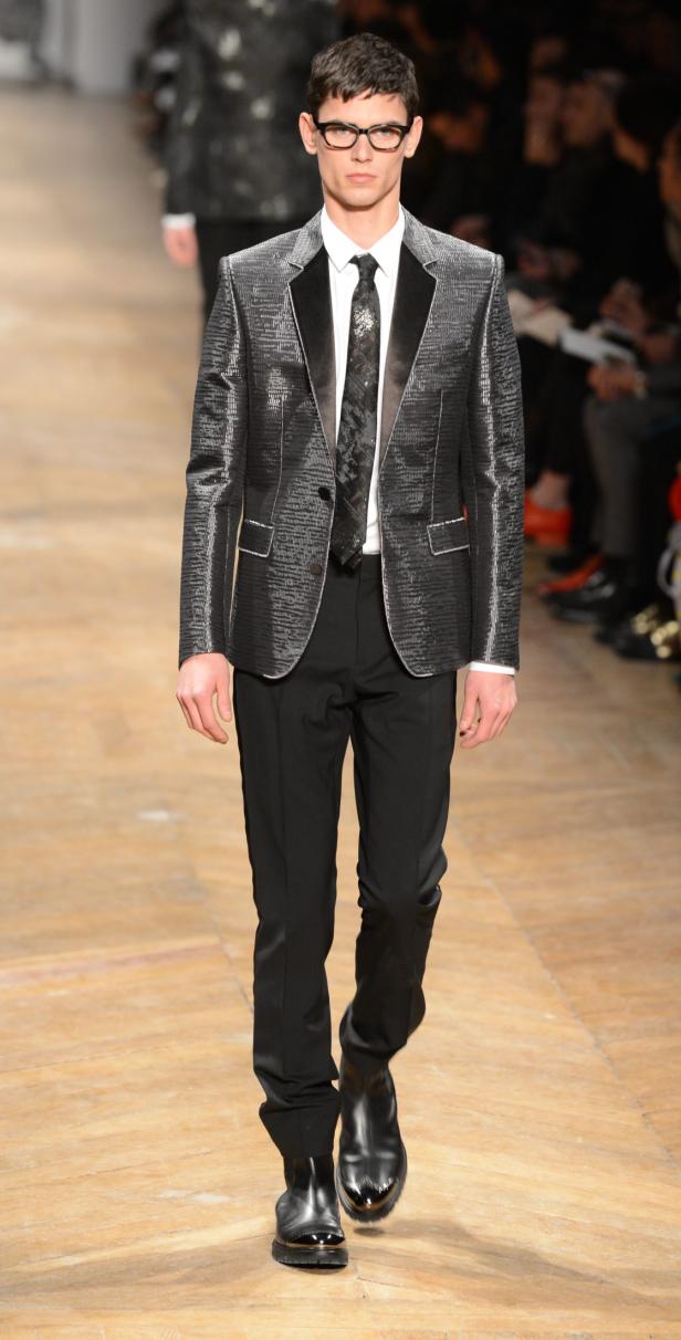 Ein Mann mit Brille trägt einen glänzenden Anzug auf einem Laufsteg.