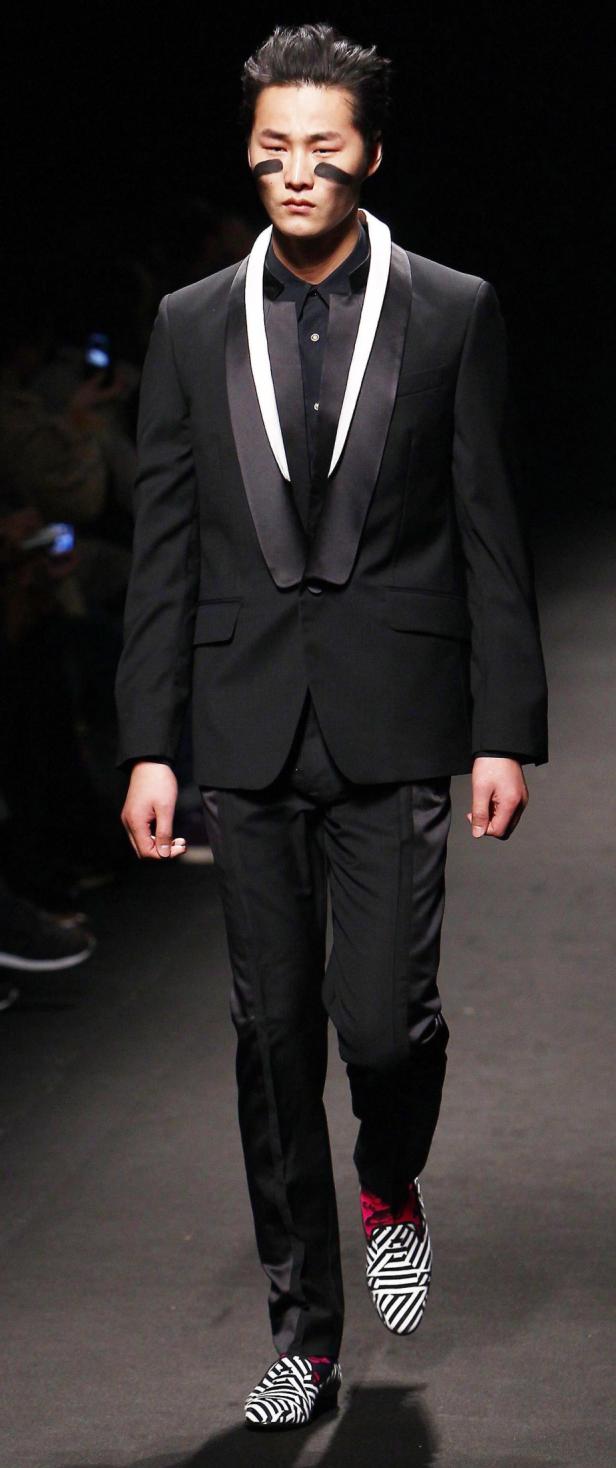 Ein Mann mit schwarzer Kriegsbemalung im Gesicht trägt einen eleganten Smoking und auffällige Schuhe.