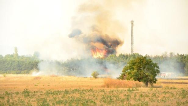Ein Waldbrand erzeugt Rauchwolken über einem Feld; im Hintergrund ist ein Sendemast zu sehen.