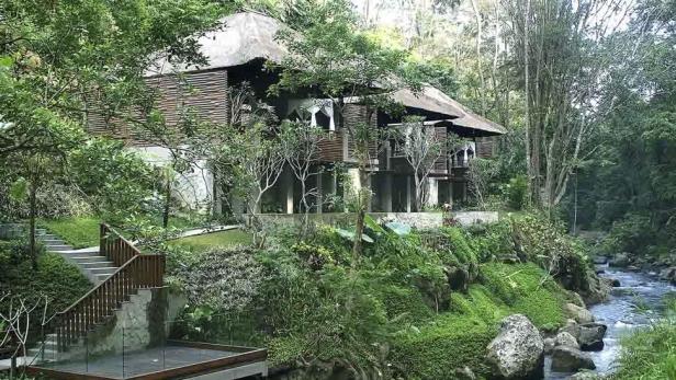 Ein zweistöckiges Haus mit Strohdach liegt inmitten üppiger Vegetation an einem Fluss.