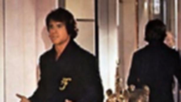 Ein Mann in einem schwarzen Bademantel mit goldenem Logo steht in einem Flur.