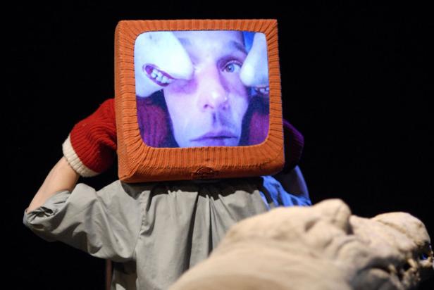 Eine Person trägt einen Fernseher als Kopfbedeckung, auf dem ein Gesicht mit verzerrten Zähnen zu sehen ist.